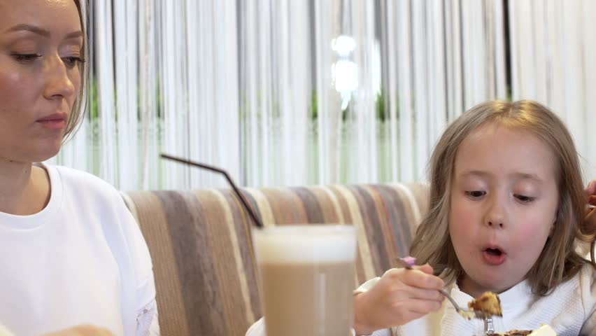 Side View - A Cute Young Mother And A Cute Little Daughter Eat Together A Delicious Dessert Sitting At A Table In A Cafe. Concept Of Joint Family Holidays