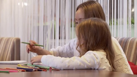 Side View - Positive Young Woman Is Drawing With Her Little Beautiful Daughter Sitting At A Cozy Table In A Cafe. Concept Of Educational Activities With Children