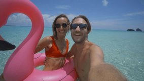Young couple take selfie portrait on idyllic beach with inflatable pink flamingo in pristine clear water in the Islands of Thailand. People travel luxury fun and cool attitude concept 