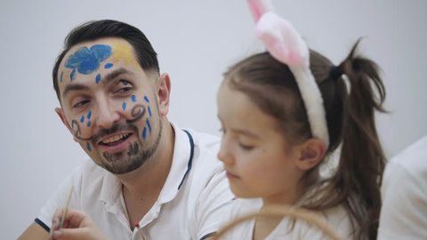 Little cute girl with bunny ears on her head is painting daddy's face in yellow and blue colors. Concept Easter holiday.