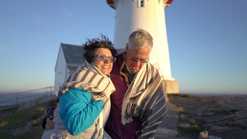 Mature couple walking at sunset on the seashore with an old lighthouse take care of each other, covering themselves with a plaid from the strong wind and hugging. Slow motion Royalty-Free Stock Footage #1026092057