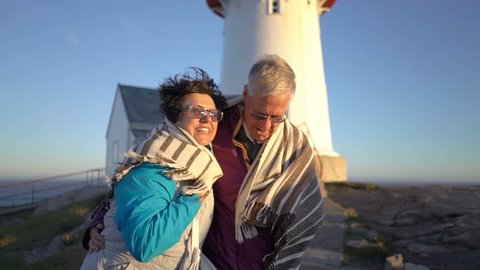 Mature couple walking at sunset on the seashore with an old lighthouse take care of each other, covering themselves with a plaid from the strong wind and hugging. Slow motion