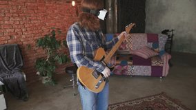 Portrait of young redhead man musician playing electric guitar in VR glasses at home, slow motion