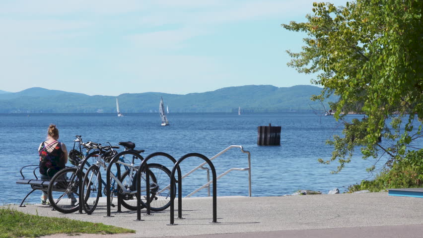 Woman sitting on the bench, taking a break next to the bike rack. Woman looking at the sailboats on the lake Champlain. Summer bliss. Wide shot.