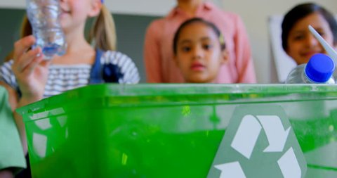 Front view of group of diverse schoolkids putting bottles in recycle container at desk in classroom. They are studying about green energy and recycle