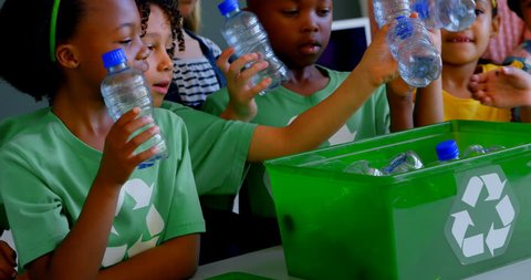 Front view of group of diverse schoolkids putting bottles in recycle container at desk in classroom. They are studying about green energy and recycle.