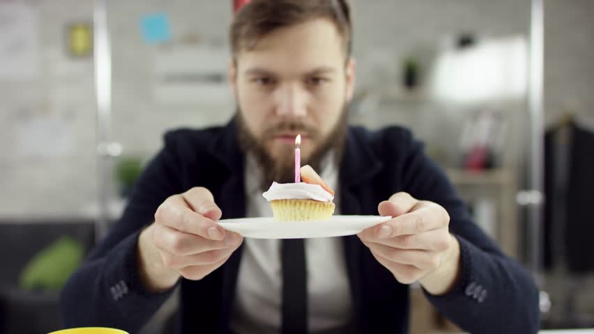 Sad bearded businessman, office worker is celebrating a lonely birthday in the office, he is blowing a candle on a small cake. He wears an office suit, white shirt and blak tie.