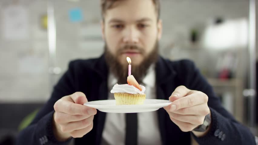 bearded businessman, office worker is celebrating a lonely birthday in the office, he is blowing a candle on a small cake. He wears an office suit, white shirt and blak tie.