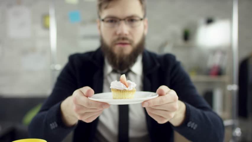 Sad working bearded businessman, office worker is celebrating a lonely birthday in the office, he is showing and giving a cup-cake. He wears an office suit, white shirt and blak tie.