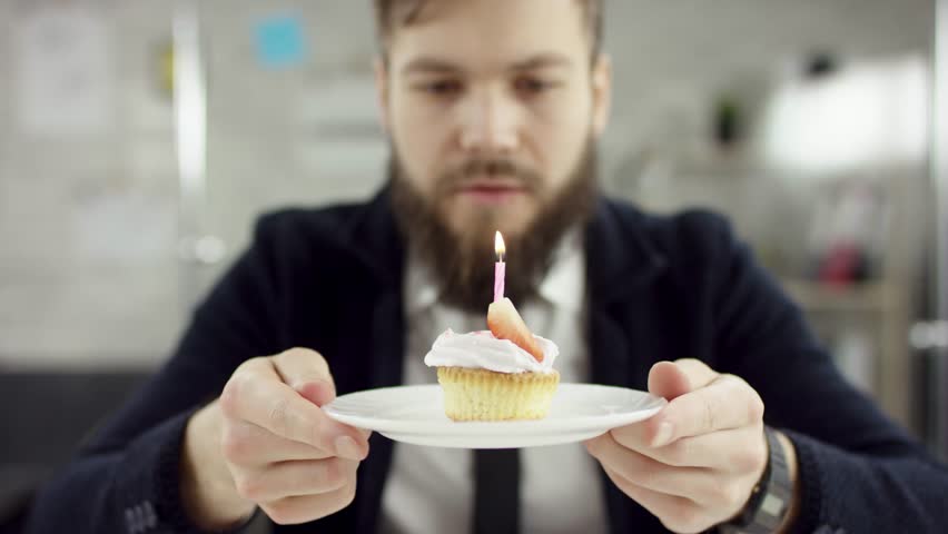 Sad serious bearded businessman, office worker is celebrating a lonely birthday in the office, he is blowing a candle on a small cake. He wears an office suit, white shirt and blak tie.