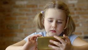 blonde preschooler girl is playing video game on smartphone and eating sandwiches in room