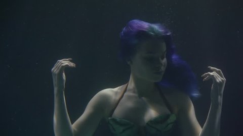 pretty young mermaid with blue hair is floating inside sea water, waving hands