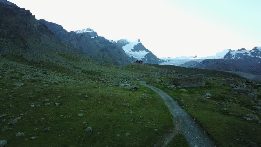 Aerial view of a hiking path winding through the green hillside towards a wooden house, with snowy rocky mountains in the background before sunrise on a summer morning, in Zermatt, Valais, Switzerland