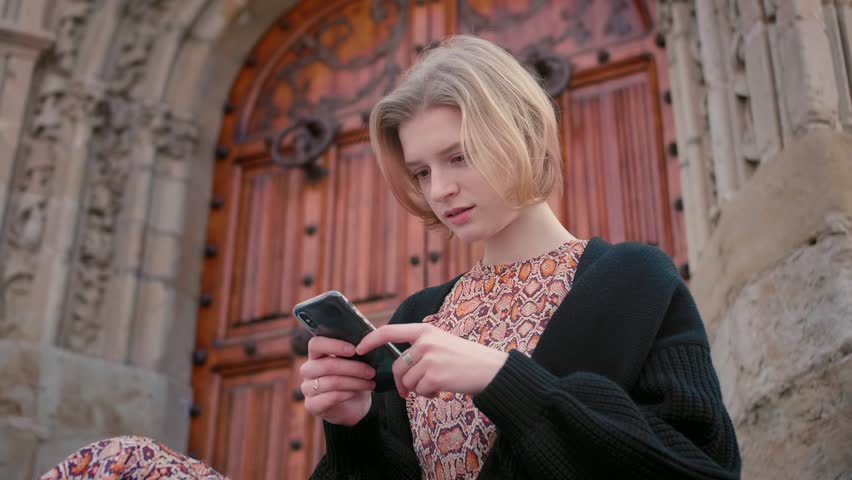 Middle plan of young blond short hair Caucasian girl having rest on city streets using smartphone communicating with friends checking mail, social networks feeling happy and cheerful