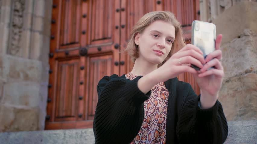 Close up of beautiful stylish Caucasian young girl with short blond hair smile and make selfie with mobile phone sitting on stairs of old European style building during summer holidays