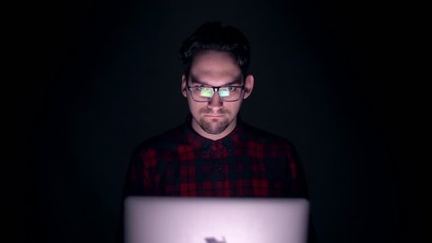 Husband watching porn sites at night. Hacker works at a laptop at night. Portrait of a smiling mad man behind the monitor in the dark. Evil laughter of the cunning genius.
