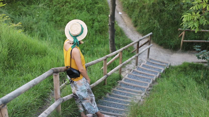 Back view of romantic woman on the wooden stairway. Stylish girl holding her hat walking down the stairs in the park.