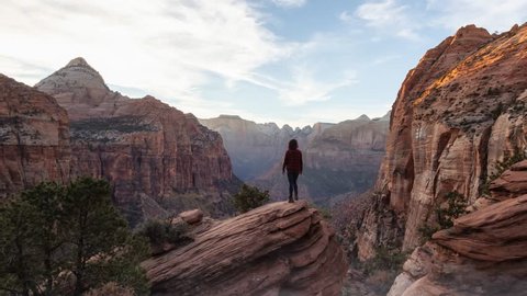 Adventurous Girl at the edge of a cliff is looking at a beautiful landscape in the Canyon during a vibrant sunset. Taken in Zion National Park, Utah, United States. Still Image Continuous Animation