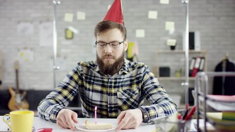 Sad attractive businessman celebrating a lonely birthday in the office, he is blowing a candle on a small cake. He is sitting in his office. Wearing formal suit, formal clothes. He is to eat the