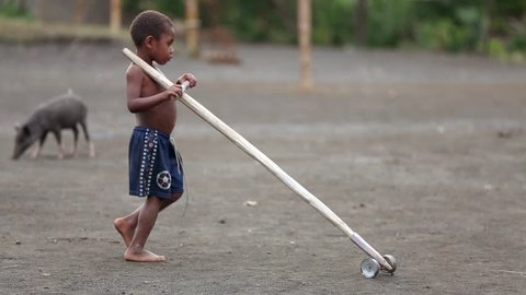 The black-skinned tribe boy is concentrated on rolling the hand-made wooden agricultural tool with two wheels. Yakel tribe kid is playing outdoor in Vanuatu village on Tanna Island, February 2015