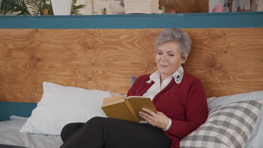 Retired female pensioner relxing in bedroom reading fiction book. Aged woman lying on stylish bed with happy face and enjoying free time. Portrait.