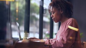 Side view of one young African college girl study indoor by the window. Black female college student typing on the laptop. urban freelancer lifestyle in the cafe. businesswoman working education clip