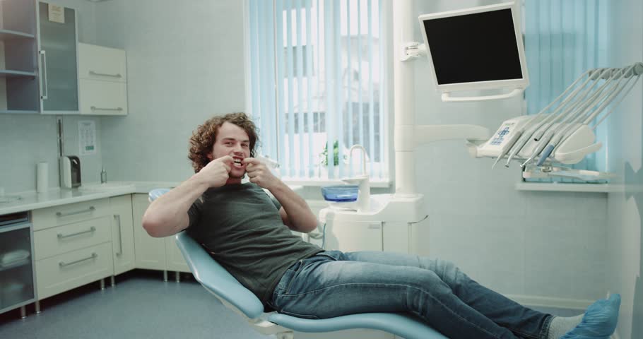 Happy patient young man at dentist sitting on the dentist chair with a big smile looking straight to the camera and showing his perfect white teeth after a oral hygiene procedure.4k