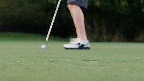 man putts the ball into the hole close up on the hole. A close-up steady video showing a man hitting a golf ball. Then a rolling golf ball on a green grassy field went into the hole.