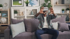 African American guy is using virtual reality glasses smiling and laughing sitting on couch and moving hands enjoying new experience. Millennials and devices concept.