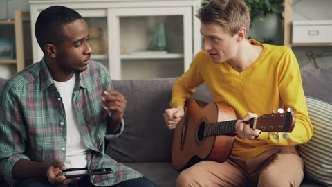 Joyful guys African American and Caucasian friends are playing the guitar, using tablet and laughing relaxing on sofa together in modern apartment. Friendship and music concept.