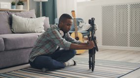 Good-looking African American blogger is adjusting camera on tripod then recording video for online vlog speaking and gesturing sitting on floor in flat.