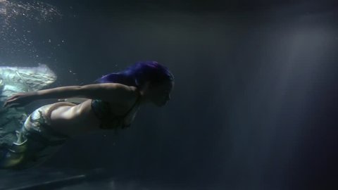 a mermaid woman with blue hair and an unusual tail swims at night under water