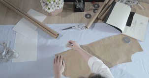 Slow motion video of the top view of the fashion designer which works at a table with curves and a pattern, draws a soap slice, around lies scissors, centimeter, a notebook with sketches, sew studio