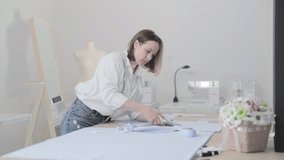 Slow motion video of the fashion designer which works at a table with curves and a pattern, she are cutting fabric, around lies scissors, centimeter, sew machine and dummy on the background