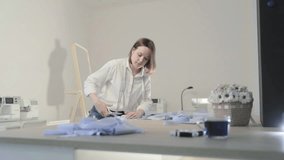 Slow motion video of the fashion designer which works at a table with curves and a pattern, she are cutting fabric, around lies scissors, centimeter, sew machine and dummy on the background