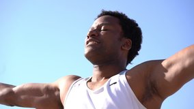 Happy african american man training at sunny day. Low angle view of smiling athletic man with closed eyes exercising against blue sky, handheld shot. Workout concept