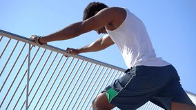 Smiling african american sportsman exercising against blue sky. Low angle view of athletic happy man in sportswear leaning at fence and working out at sunny day, handheld shot. Workout concept
