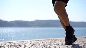 Low section if man in shorts and sneakers running at riverside. Cropped shot of mature athlete in sportswear jogging along embankment at sunny day, slow motion, tracking shot. Working out concept
