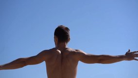 Muscular shirtless man stretching arms against blue sky. Low angle view of athletic bare-chested man exercising outdoor at sunny day, back view. Working out concept