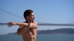 Muscular shirtless man stretching arms outdoor. Athletic handsome young man exercising and doing stretching exercise at sunny day, handheld shot. Working out concept
