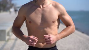 Cropped shot of shirtless sportsman running at riverside. Mid section of muscular bare-chested man jogging along embankment at sunny day, tracking shot. Workout concept