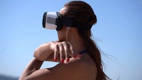 Girl in vr headset stretching against blue sky. Young woman in earphones and virtual reality headset exercising outdoor at sunny day, handheld shot. Technology concept