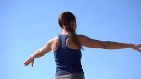 Back view of sporty girl stretching arms outdoor. Rear view of athletic young woman in sportswear and earphones exercising against blue sky at sunny day, low angle view. Workout concept