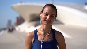 Young woman in earphones smiling at camera on street. Portrait of beautiful sporty girl listening music and looking at camera while standing outdoor at sunny day. Active lifestyle concept