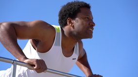 Smiling sporty man training on railing against blue sky. Low angle view of muscular african american athlete doing push ups at sunny day. Working out concept