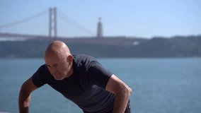 Concentrated mature man doing push ups at riverside. Slider shot of focused bald athlete in t-shirt working out near river at sunny day. Active lifestyle concept