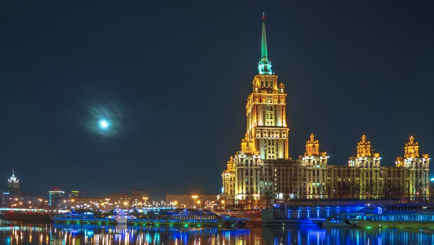 Hotel Ukraine illuminated, mirrored in Moscow river, on a calm spring night. Moon moving across the sky. Passenger pleasure boats at the pier. Time lapse of Moscow at night
