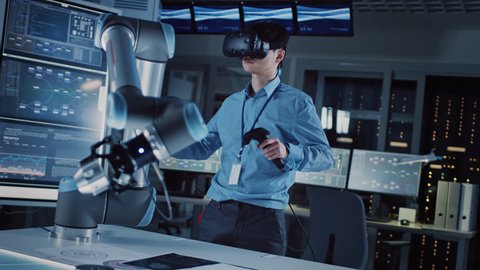 Professional Japanese Development Engineer in Blue Shirt is Controlling a Futuristic Robotic Arm with a Virtual Reality Headset and Joysticks in a High Tech Research Laboratory with Modern Equipment.
