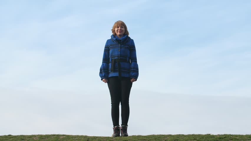  Hilarious view of smiling blonde woman in a striped blue jacket and leggings jumping up and making Saint Andrew Cross outdoors in spring in slo-mo