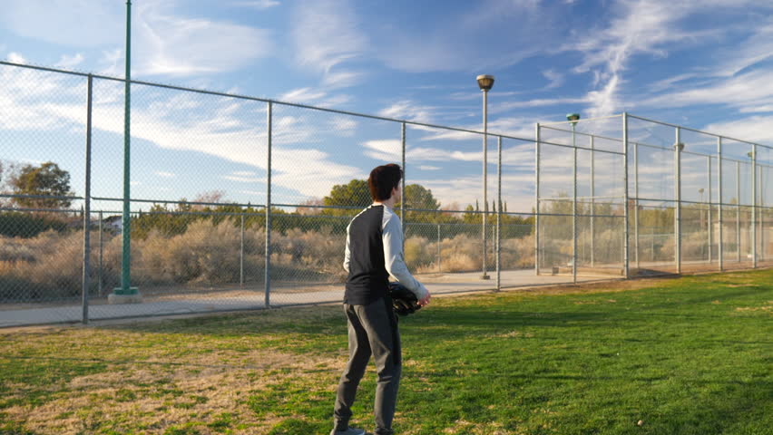 Two young athletic men playing catch together with a glove in a grassy park baseball field under blue skies during sunset.